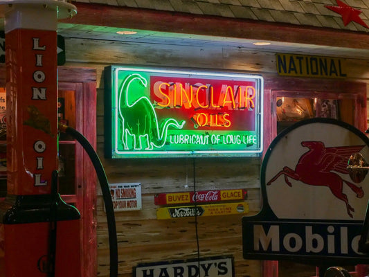Glowing vintage Sinclair Oils neon sign with green dinosaur and red lettering, mounted on a rustic wooden wall among other classic gas and oil memorabilia including a Mobil Pegasus sign and Lion Oil pump.