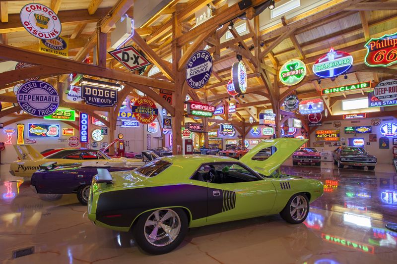 Finished garage with polished floors and multiple vintage Mopar muscle cars including a Dodge Superbird and Challenger under bright neon lighting with tons of porcelain signs hanging from ceiling and neon on walls