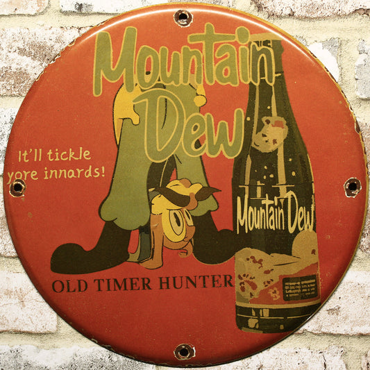 Vintage Mountain Dew Old Timer Hunter porcelain sign – 12 inch round with hillbilly character and bottle, slogan "It’ll tickle yore innards!" on red background, mounted on brick wall.