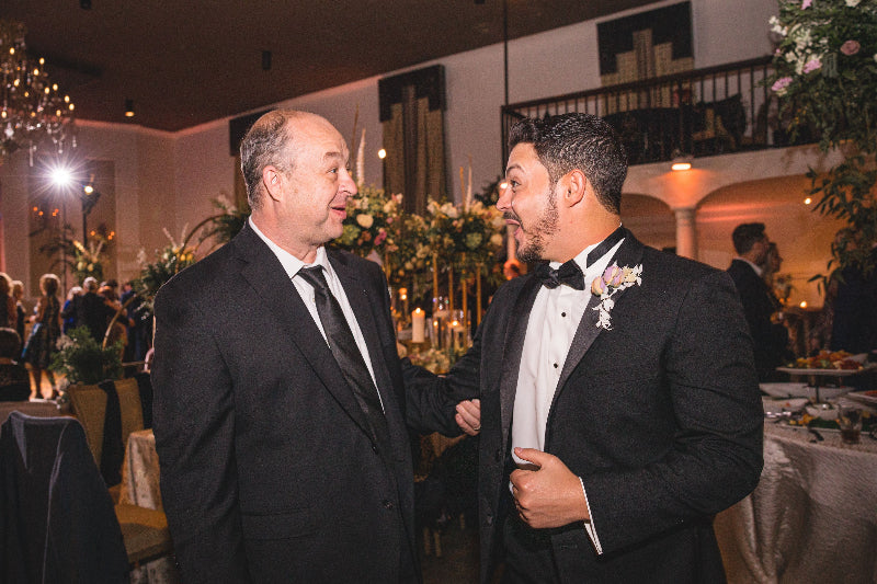 Two men in formal attire shaking hands at an event with tables and decorations in the background.